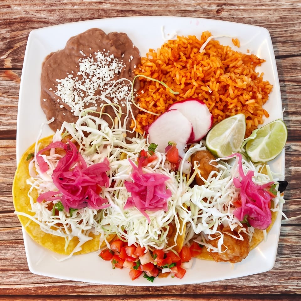 Three fish tacos with pickled onions, cabbage, rice, beans, and lime on a white plate.