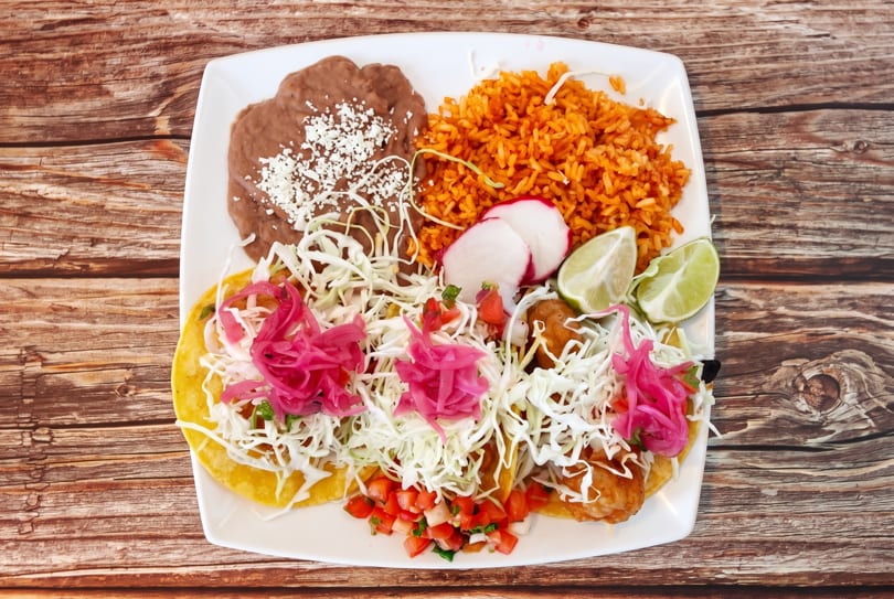 Three fish tacos with pickled onions, cabbage, rice, beans, and lime on a white plate.