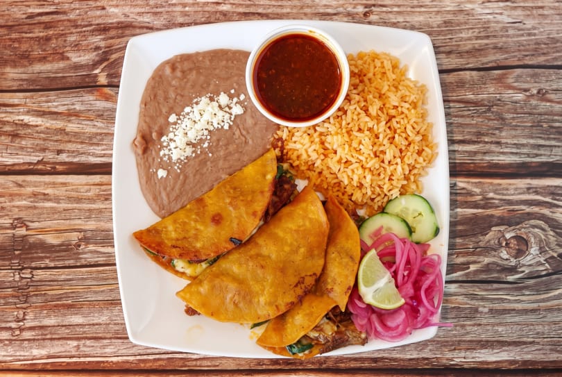 Three golden-fried tacos, rice, refried beans, and a side of red sauce on a white plate.