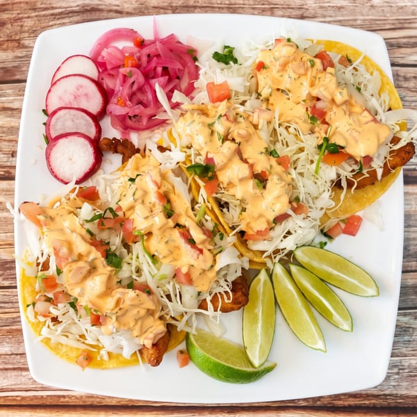 Three fish tacos with cabbage, pico de gallo, and creamy sauce, served with pickled onions, radishes, and lime wedges.