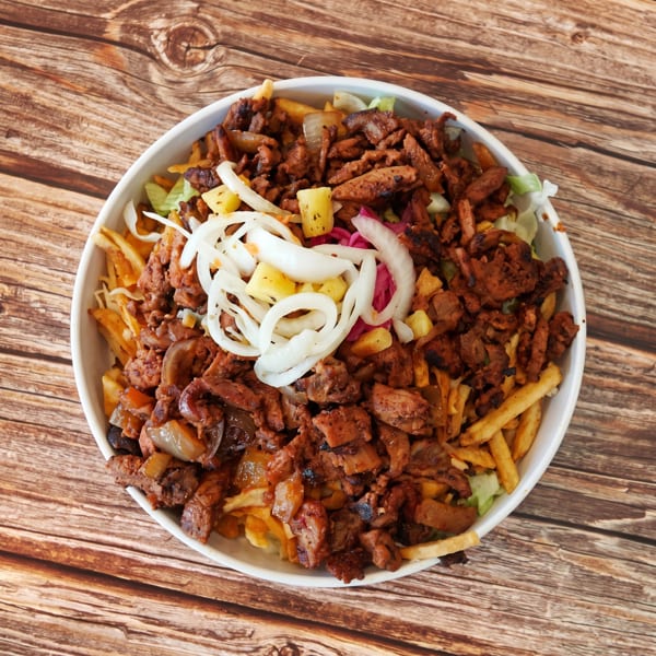 A white bowl filled with fries, grilled meat, lettuce, pineapple, and sliced onions on a wooden table.