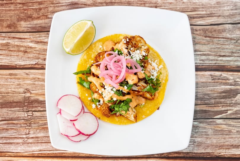 A cauliflower taco with pickled onions, cotija cheese, and sauce, served with lime and radishes.