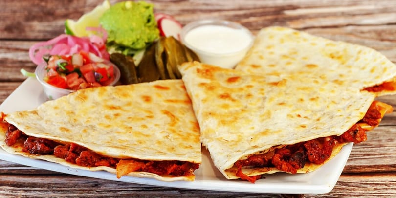 Three quesadillas with red meat filling, served with guacamole, salsa, sour cream, and pickled vegetables.